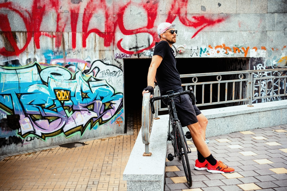 A Bicycle Rider and his Bicycle Leaning on a Railing While Wearing AirPods, Shades, and a Pair of Red Cycling Shoes